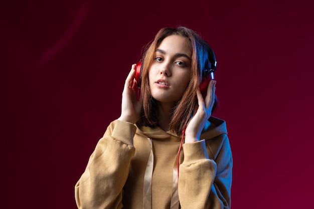 Joven hermosa chica hipster de moda vestida con una sudadera con capucha escuchando música en auriculares rojos en un fondo dramático rojo de estudio