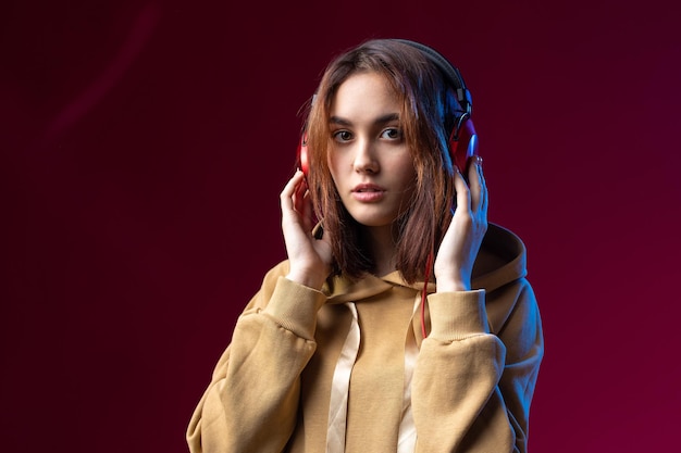 Joven hermosa chica hipster de moda vestida con una sudadera con capucha escuchando música en auriculares rojos en un fondo dramático rojo de estudio