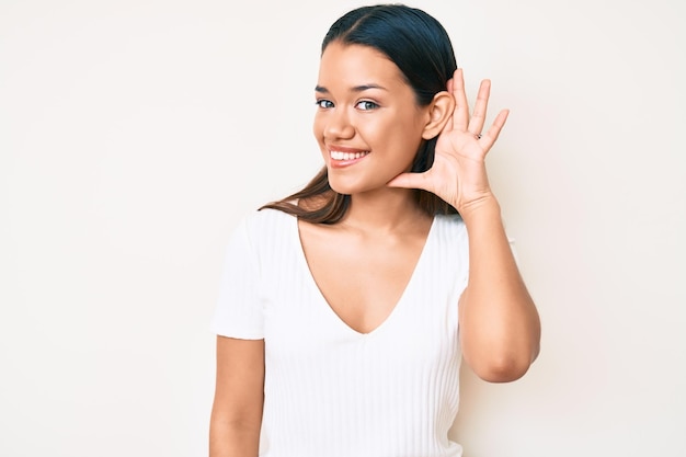 Foto joven y hermosa chica latina con una camiseta blanca casual sonriendo con la mano sobre la oreja escuchando y escuchando rumores o chismes concepto de sordera