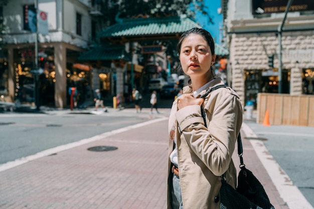 Joven hermosa chica linda con abrigo elegante con bolso de moda bajo el sol. concepto de estilo de vida local de dama encantadora. mujer bonita y pensativa parada en la calle de un día soleado y mirando al frente pensativa.