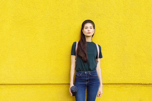 Joven hermosa chica morena en jeans una camiseta verde cintas para el cabello y una mochila rosa posando sobre un muro de hormigón amarillo