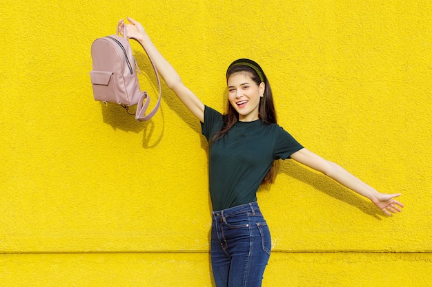 Joven hermosa chica morena en jeans una camiseta verde cintas para el cabello y una mochila rosa posando sobre un muro de hormigón amarillo