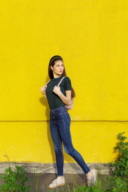 Joven hermosa chica morena en jeans una camiseta verde cintas para el cabello y una mochila rosa posando sobre un muro de hormigón amarillo