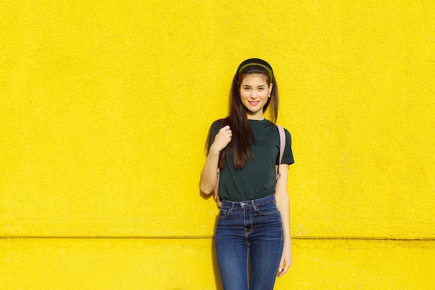 Joven hermosa chica morena en jeans una camiseta verde cintas para el cabello y una mochila rosa posando sobre un muro de hormigón amarillo