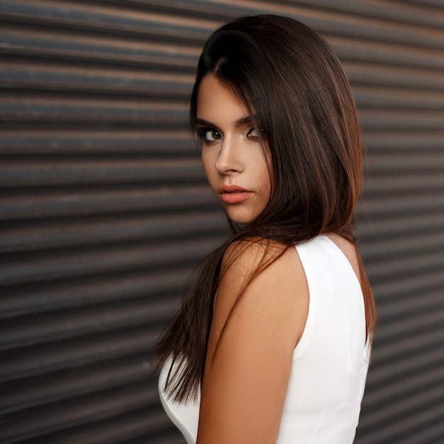 Joven hermosa chica morena con maquillaje en vestido blanco cerca de persianas