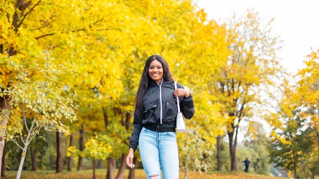 Joven hermosa chica negra feliz en ropa casual de moda mira con bolso de moda camina en el parque con follaje otoñal dorado brillante