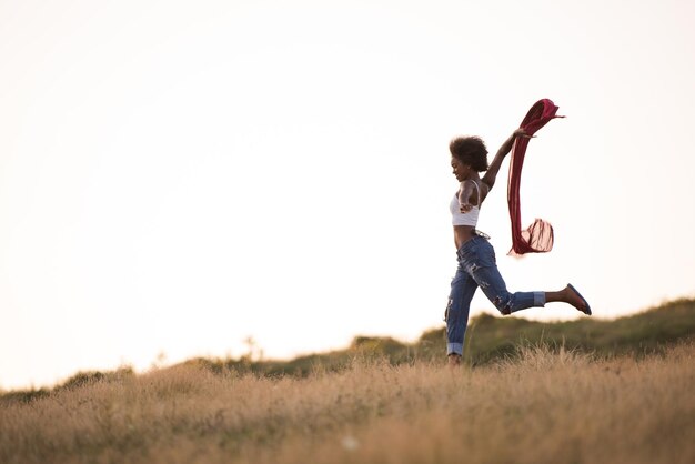 Joven hermosa chica negra se ríe y baila al aire libre con una bufanda en las manos en un prado durante la puesta de sol