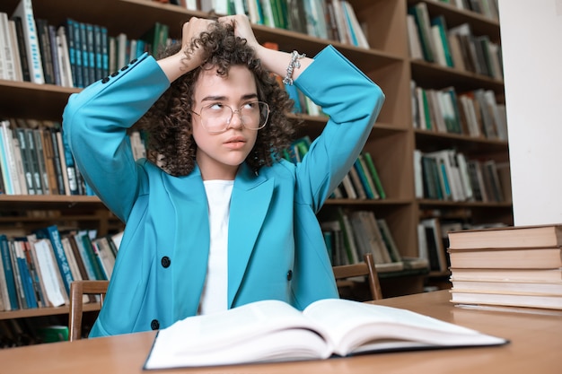 Joven hermosa chica rizada en gafas sentado en la biblioteca