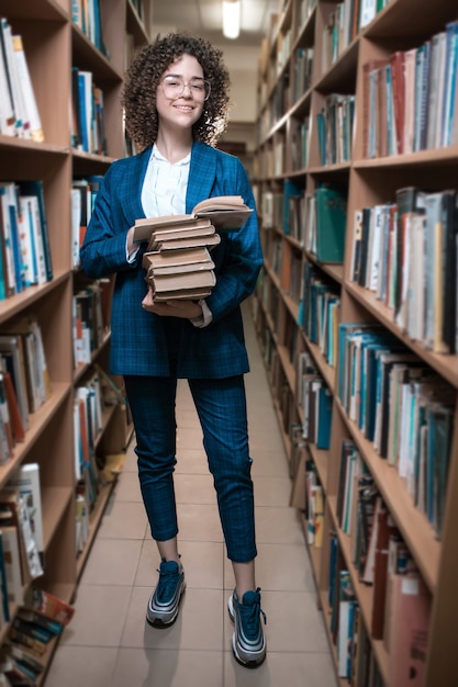 Joven hermosa chica rizada en gafas y un traje azul está de pie en la biblioteca