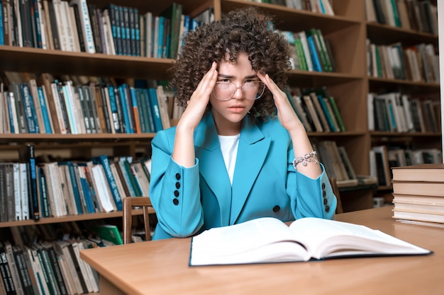 Joven hermosa chica rizada en gafas y traje azul sentado con libros en la biblioteca.