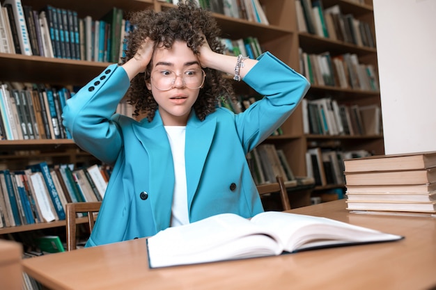 Joven hermosa chica rizada en gafas y traje azul sentado con libros en la biblioteca.