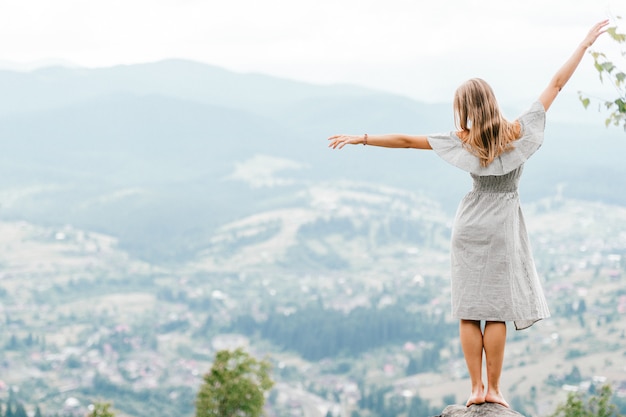 Joven hermosa chica rubia descalza con el pelo largo en vestido de verano de pie en la cima de la montaña conquistada