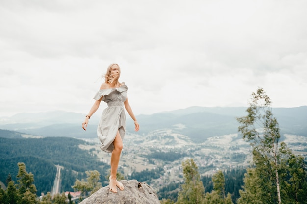 Joven hermosa chica rubia descalza con el pelo largo en vestido de verano de pie en la cima de la montaña