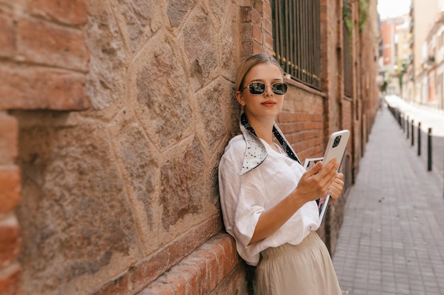 Una joven y hermosa chica rubia con gafas de sol oscuras posa cerca de una pared de piedra con una blusa blanca y un