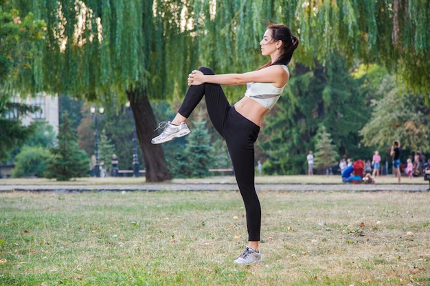 Joven hermosa chica sexy feliz se estira en el jardín del parque. antecedentes de la gente. con cabello oscuro y traje deportivo blanco y negro y felicidad y sonrisa. en temporada de verano u otoño.