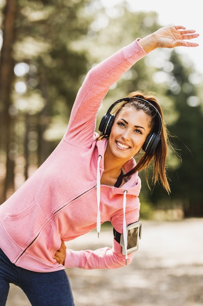 Foto joven hermosa corredora sonriente escuchando música mientras se estira antes de trotar en la naturaleza.