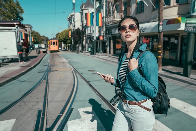 joven y hermosa dama viajera con gafas de sol sosteniendo un celular buscando dirección en el mapa en línea en el área de la ciudad de Castro. lugar de un gran grupo de cultura gay lesbiana LGBT. chica cruza la calle en el tranvía