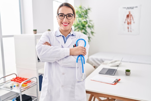 Joven hermosa doctora hispana sonriendo confiada de pie con gesto de brazos cruzados en la clínica