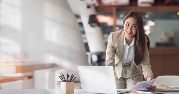 Joven hermosa y encantadora mujer de negocios asiática sonriendo y mirando a la cámara mientras trabaja en una computadora portátil y documenta en la oficina