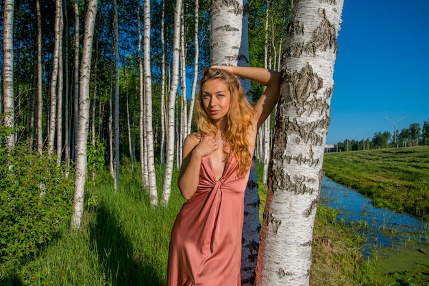 Joven hermosa esbelta en un vestido largo de coral posando en un bosque de abedules sonriendo y pecho desnudo