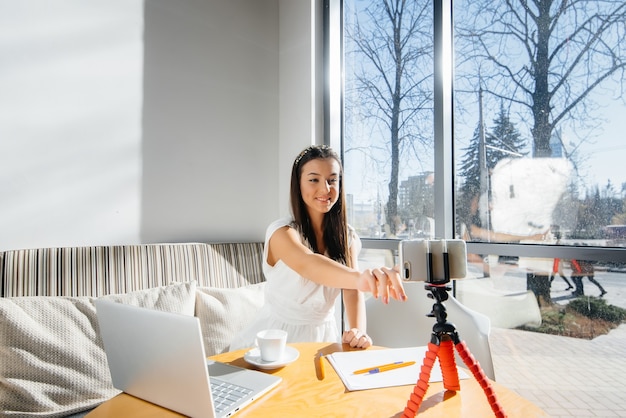 Una joven hermosa está sentada en un café, grabando blogs de video y charlando en las redes sociales.