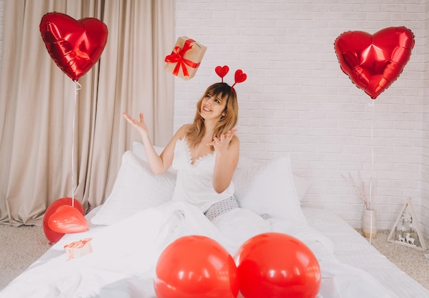 Una joven hermosa y feliz lanza un regalo al aire mientras está sentada en la cama. día de San Valentín