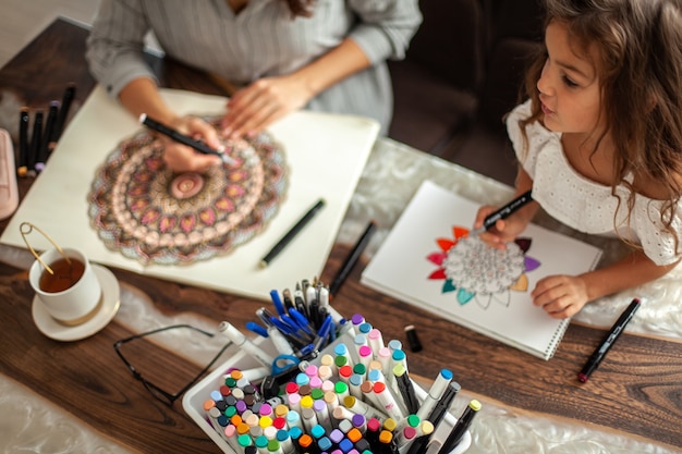 Joven hermosa madre e hija linda dibujan un patrón de mandala juntos marcadores artísticos y colore ...