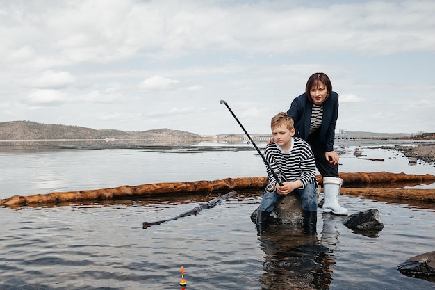Joven, hermosa madre e hijo se divierten pescando en el lago. Mamá e hijo con chalecos a rayas en la orilla del río con una caña de pescar.