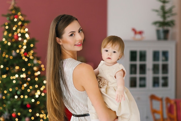 Una joven hermosa madre sostiene a su hija de un año con un vestido beige sobre el fondo de árboles de Navidad en la sala de estar de la casa