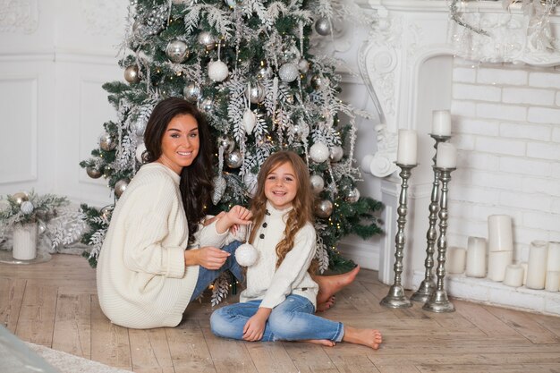 Joven hermosa madre y su pequeña hija linda en suéteres acogedores blancos y una manta se divierten y se abrazan bajo un árbol de Navidad decorado. Interior clásico hogar festivo.