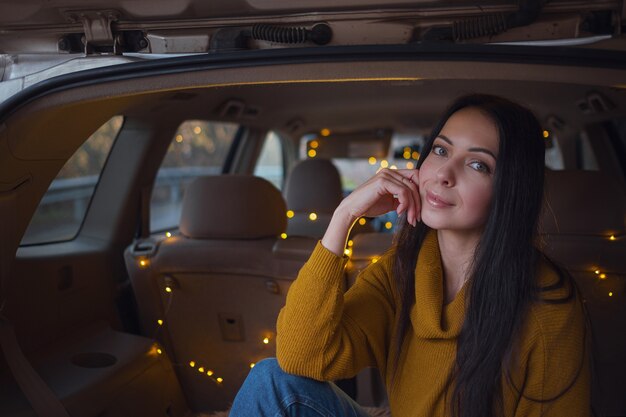 Joven hermosa morena disfruta relajándose en el maletero de su coche. Una guirnalda amarilla y una tela escocesa beige crean comodidad.