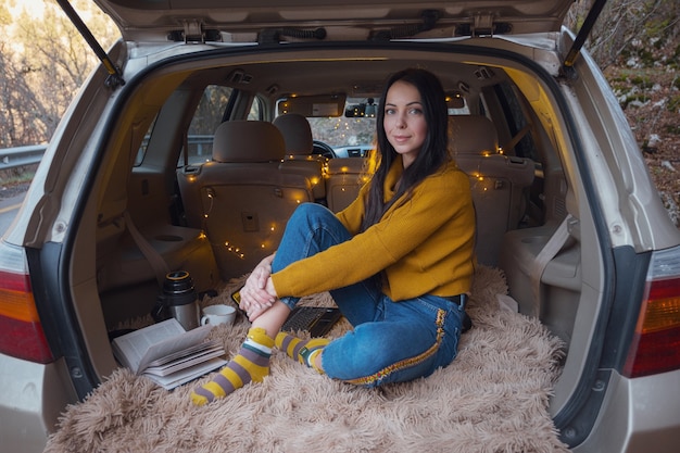 Joven hermosa morena disfruta relajándose en el maletero de su coche. Una guirnalda amarilla y una tela escocesa beige crean comodidad.