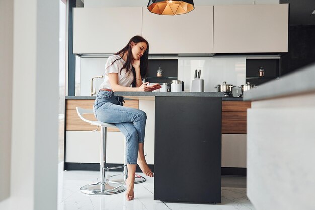 Una joven y hermosa morena con ropa informal se sienta en la cocina con una bebida fresca a la mañana