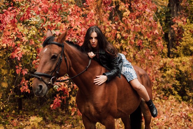 Joven hermosa morena con un vestido y una chaqueta de cuero negro es sexy acostada sobre un caballo