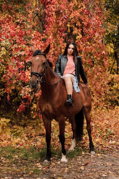 Joven hermosa morena en un vestido y chaqueta de cuero negro sexy se sienta en un caballo