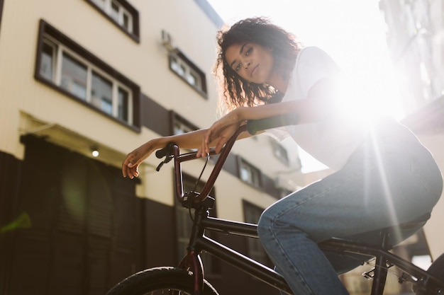 Joven hermosa mujer afroamericana con cabello rizado oscuro en camiseta blanca y jeans mirando soñadoramente a la cámara mientras monta en bicicleta al aire libre