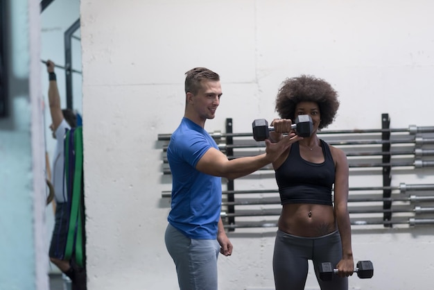 joven y hermosa mujer afroamericana haciendo flexiones de bíceps con entrenador de fitness en un gimnasio