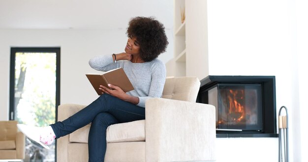 Joven hermosa mujer afroamericana sentada frente a la chimenea en casa en un frío día de otoño y leyendo un libro