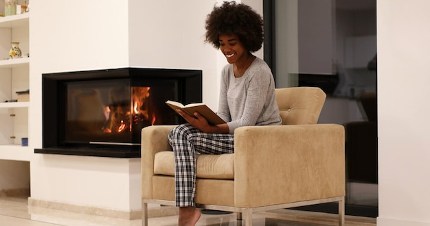 Joven hermosa mujer afroamericana sentada frente a la chimenea en casa en un frío día de otoño y leyendo un libro