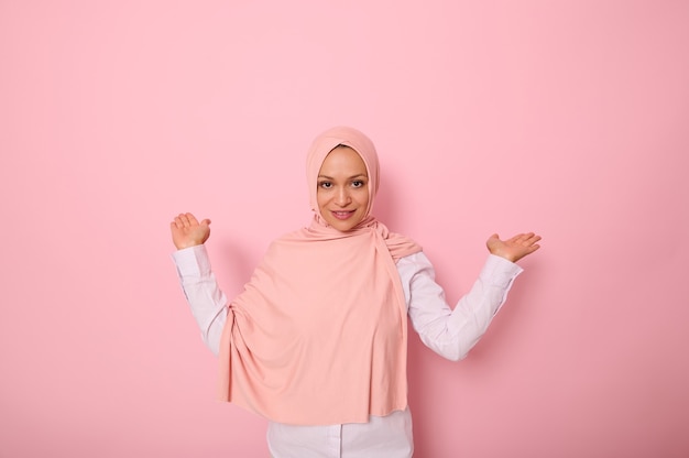 Joven hermosa mujer árabe musulmana en hijab rosa sobre fondo rosa aislado sosteniendo un espacio de copia imaginario en la palma para insertar una publicidad