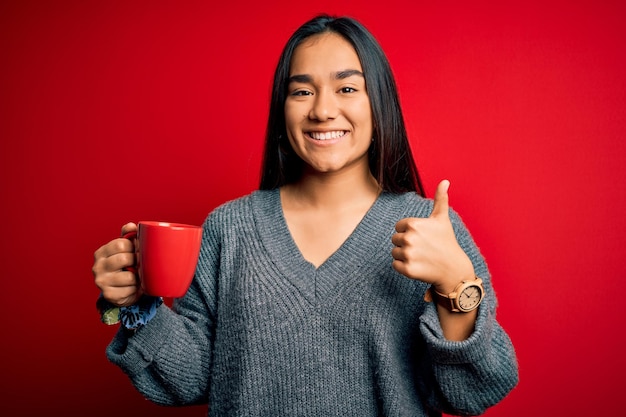 Joven hermosa mujer asiática bebiendo una taza de café de pie sobre un fondo rojo aislado feliz con una gran sonrisa haciendo bien firmar el pulgar hacia arriba con los dedos excelente señal