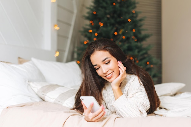 Joven hermosa mujer asiática con cabello largo oscuro en un acogedor suéter de punto blanco usando un teléfono inteligente móvil en la cama en la habitación con árbol de Navidad