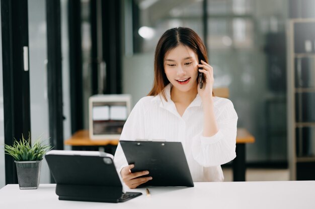 Joven y hermosa mujer asiática escribiendo en una tableta y una computadora portátil mientras está sentada en la oficina de la mesa blanca de trabajo