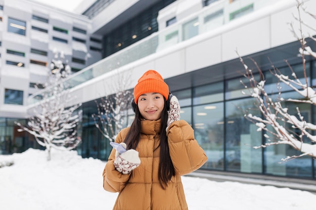 Joven hermosa mujer asiática escucha música con auriculares mientras camina en un día nevado de invierno usa una aplicación en línea para el teléfono