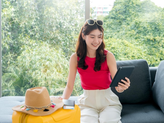 Joven hermosa mujer asiática feliz con gafas de sol usando una tableta digital con boleto de avión de pasaporte y sombrero de playa en una maleta amarilla sentada en el sofá lista para viajar en concepto de vacaciones de verano