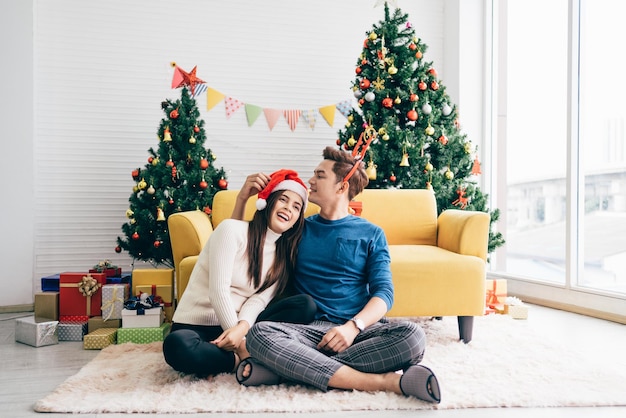 Joven hermosa mujer asiática feliz con un sombrero de Santa Claus sentada junto a su novio en casa con un árbol de Navidad en el fondo Imagen con espacio de copia