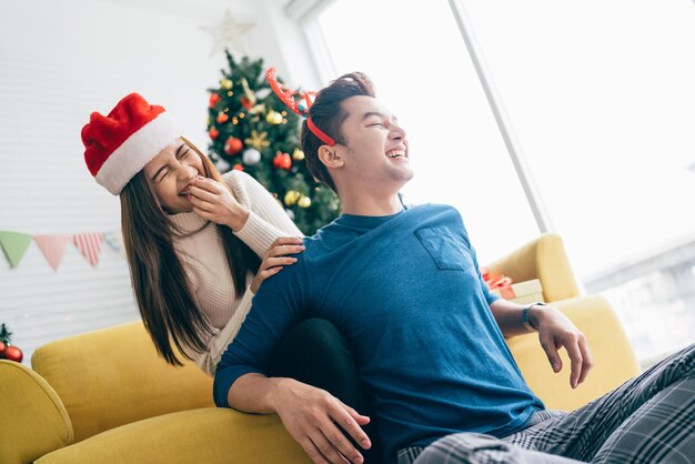 Joven hermosa mujer asiática feliz con sombrero de Santa Claus sorprende y se ríe en la parte de atrás de su novio en casa con un árbol de Navidad en el fondo Imagen con espacio de copia