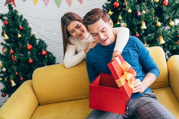 Joven hermosa mujer asiática feliz sorprende a su novio con un regalo de Navidad en casa con un árbol de Navidad en el fondo Imagen con espacio de copia