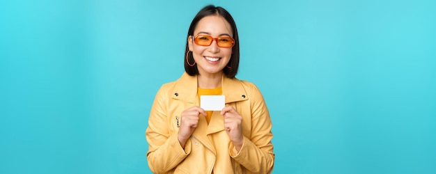 Joven hermosa mujer asiática mostrando tarjeta de crédito sonriendo eligiendo banco de pie sobre fondo azul Copiar espacio