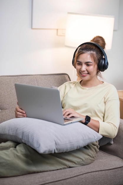 Joven hermosa mujer asiática relajándose y escuchando música usando auriculares ella está acostada en el sofá Mujer joven disfrutando en casa usando computadora portátil
